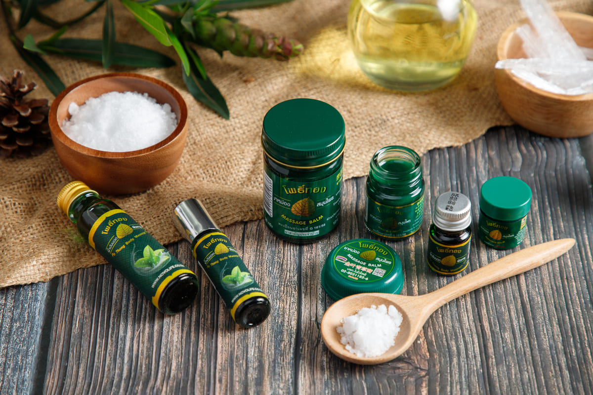 Thai herbal wellness lifestyle showing Pho Tong Green Cooling Balm 50g glass jar used for muscle pain relief, headache therapy, and travel first aid kit essentials